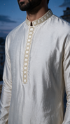 A close-up of a man wearing a cream-colored kurta with gold chain-like embroidery down the front placket and around the collar, set against a soft blue sky background.