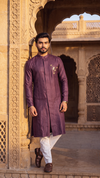 A man wearing a long-sleeved, knee-length plum purple kurta with vertical pleats and a small gold embroidered motif on the chest, paired with white trousers and brown shoes, standing in front of a historic sandstone archway.