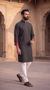 A man wearing a dark gray patterned kurta with red cuffs, white trousers, and brown sandals, walking in front of a historic sandstone building with arches.