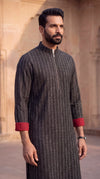 A man wearing a dark charcoal gray textured long kurta with a half-zip and red contrast cuffs, standing in front of a warm beige stone architectural background.