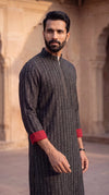 A man wearing a dark gray textured long kurta with red cuff lining, standing in front of a warm beige stone architectural background.