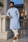 A man wearing a light blue long kurta with vertical gold embroidery, white trousers, and brown loafers, posing in an elegant outdoor courtyard with classical columns.