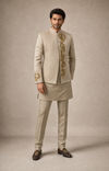 A man modeling a traditional Indian ensemble featuring a beige textured indowestern  jacket with gold embroidery, a matching beige kurta, beige trousers, and dark brown loafers, set against a neutral beige studio backdrop.