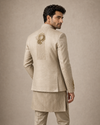 A man wearing a light beige traditional Indian suit with a textured kurta and matching trousers, shown from the back. The jacket features ornate gold embroidery on the upper back and sleeve cuffs, set against a neutral beige studio background.
