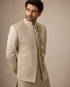 A man wearing a beige textured indowestern jacket with ornate gold embroidery on the front panel and cuffs, paired with a matching beige kurta, set against a neutral beige background.