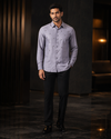 A man wearing a light lavender-gray long-sleeve button-up shirt with small embroidered details, paired with black trousers and black dress shoes, standing in a dark, modern interior setting.
