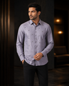 A man wearing a light lavender-gray long-sleeve button-up shirt with small embroidered details, paired with black trousers, standing in a dimly lit modern interior.