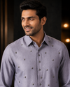 A man wearing a light lavender-gray long-sleeve button-up shirt with small metallic decorative embellishments, shown in a close-up portrait against a dark, softly lit background.