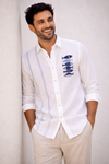 A man wearing a white long-sleeve linen shirt with blue vertical stripes and a stack of fish embroidery on the chest, paired with beige trousers, leaning against a light wall.