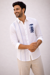 A man wearing a white long-sleeve linen shirt with blue embroidered fish on the chest and vertical blue stripes, paired with beige trousers, posing against background.