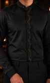 A close-up of a man wearing a black long-sleeve shirt with a stand collar and a distinctive wavy vertical line of gold-toned embellishments down the front placket, paired with black trousers.