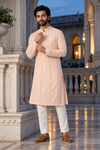 A man wearing a light peach-pink long kurta with white trousers and brown shoes, standing in a grand, elegant courtyard with classical architecture and warm lighting.