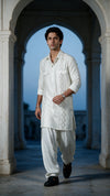 A man wearing a white embroidered kurta and matching pants walks through a stone archway corridor at dusk.