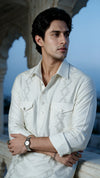 A man wearing a white long-sleeve button-up shirt with light gray embroidered geometric patterns on the chest, posed outdoors against a stone archway and blue sky background.
