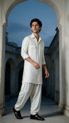 A man wearing a traditional white embroidered kurta and matching loose trousers, standing in a stone courtyard framed by arches under a blue sky.