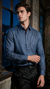 A man wearing a blue long-sleeve button-up shirt with black geometric embroidered patterns on the chest and sleeves, paired with black pants, standing in front of a dark window.