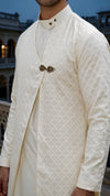 A close-up of a man wearing a cream-colored, long-sleeved traditional kurta with a subtle woven geometric pattern and a decorative metallic chest pin, set against a blurred architectural background at dusk.