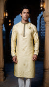 A man wearing a pale yellow embroidered kurta with white trousers, standing in a traditional architectural setting with stone pillars and hanging lanterns.