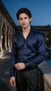 A man wearing a dark navy blue satin long-sleeve shirt with intricate embroidered detailing on the chest and cuffs, paired with black trousers, standing outdoors in front of a classical building at dusk.