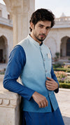 A man wearing a light blue quilted waistcoat over a royal blue kurta, standing in a courtyard with stone pillars and arches in the background.