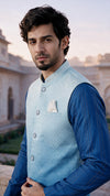 A man wearing a light blue quilted jacket over a royal blue long-sleeve shirt, standing outdoors in front of a soft-focus architectural background.