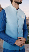 A man wearing a light blue quilted waistcoat over a royal blue long-sleeve kurta, standing outdoors with his hands clasped in front of a softly blurred architectural background.