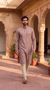 A man wearing a light brown long-sleeve kurta with subtle white embroidery and cream trousers, walking in a sunlit courtyard with sandstone arches and potted plants.