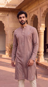 A man wearing a dusty rose long-sleeve kurta with subtle white embroidery and cream trousers, standing in front of a warm sandstone courtyard with arches.