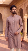 A man wearing a dusty rose pink kurta with subtle white embroidery, standing in front of a traditional sandstone building with arches.