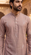 A man wearing a light brownish-pink long-sleeve kurta with white embroidered paisley patterns, standing in front of a warm-toned stone building.