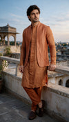 A man wearing a traditional Indian outfit in rust-orange and brown stands on a rooftop terrace at dusk, leaning on a stone railing with a cityscape and pavilion in the background.