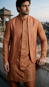A man wearing a traditional Indian outfit consisting of a rust-orange textured waistcoat, a matching striped kurta, and trousers, standing outdoors against a blurred architectural background.