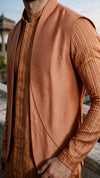 A close-up of a man wearing a sleeveless textured vest over a matching orange and brown striped kurta, photographed outdoors with a blurred architectural background.
