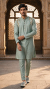 A man wearing a light sage green kurta and matching trousers with gold embroidery on the shoulders, walking in front of a traditional stone archway.