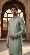A man wearing a light sage green kurta with gold embroidery on the chest, standing in front of an ornate stone archway.