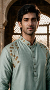 A man wearing a light sage green kurta with gold floral embroidery on the shoulders, standing in front of an ornate stone archway.