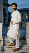 A man wearing a cream-colored Indian kurta and matching trousers with brown leather shoes, standing on stone steps in front of a historic building at dusk.