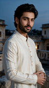 A man wearing a cream-colored embroidered kurta stands outdoors against a blurred evening cityscape background.