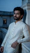 A man wearing a cream-colored embroidered kurta leans against a stone pillar outdoors at dusk, with a softly blurred architectural background.