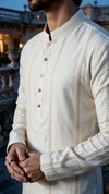 A man wearing a cream-colored long-sleeve kurta with vertical embroidered details and gold buttons, photographed outdoors against a blurred architectural background.