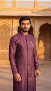 A man wearing a long-sleeved, deep purple kurta with gold embroidery on the chest, standing in front of a warm, sunlit sandstone architectural background.