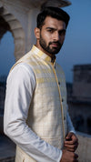 A man wearing a cream and gold checkered waistcoat over a white long-sleeve shirt, standing outdoors against a soft-focus architectural background at dusk.