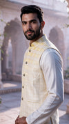 A man wearing a light yellow ombre checkered waistcoat over a white kurta, standing in front of a softly blurred traditional stone building with arches.