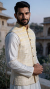 A man wearing a cream and light yellow checkered waistcoat over a white kurta, standing outdoors in front of a blurred stone building.