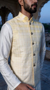 A man wearing a light yellow ombre
checkered jacket vest over a white kurta, standing outdoors in front of a stone archway.
