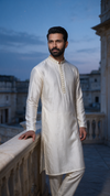 A man wearing a cream-colored Indian kurta and matching trousers stands on a balcony at dusk, with a stone building and blue sky in the background.